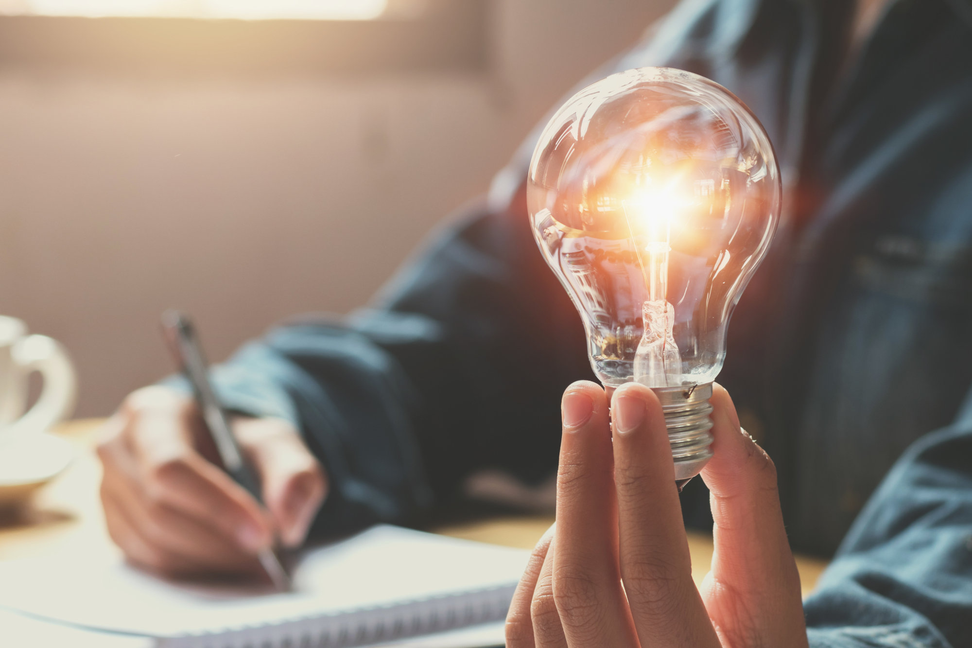 Man holding lit light bulb while writing on a notepad